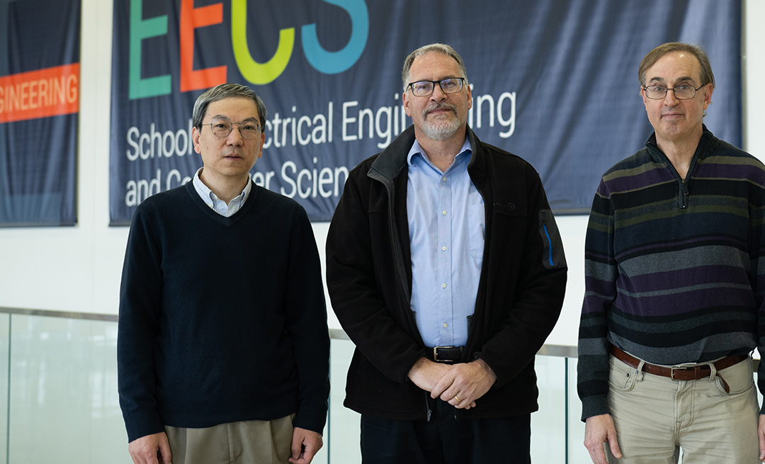 Three people pose outside in front of an electrical engineering and computer science banner